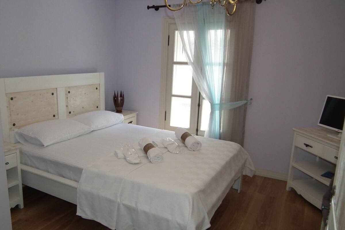A neatly made bed with white linens and towels, a bedside table with a lamp, and a television on a stand.