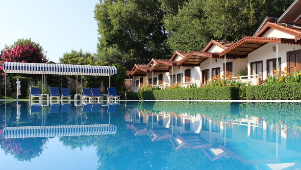 A swimming pool with lounge chairs, a covered seating area, and a row of villas with balconies. - Hulusi Hotel (Manavgat)