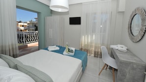A hotel room with a bed, a desk, a chair, a TV, and a balcony with a view of buildings. - Dionisos Elia (Nea Propontida)