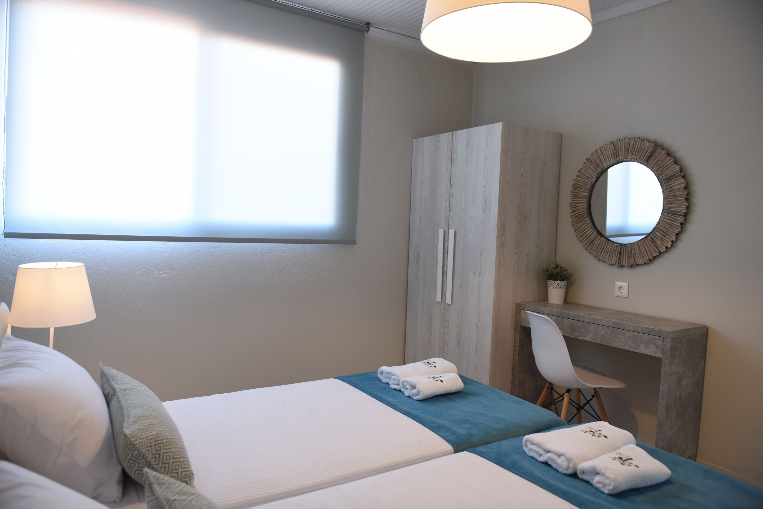 A hotel room with two beds, a desk, a chair, a mirror, and a window with blinds.