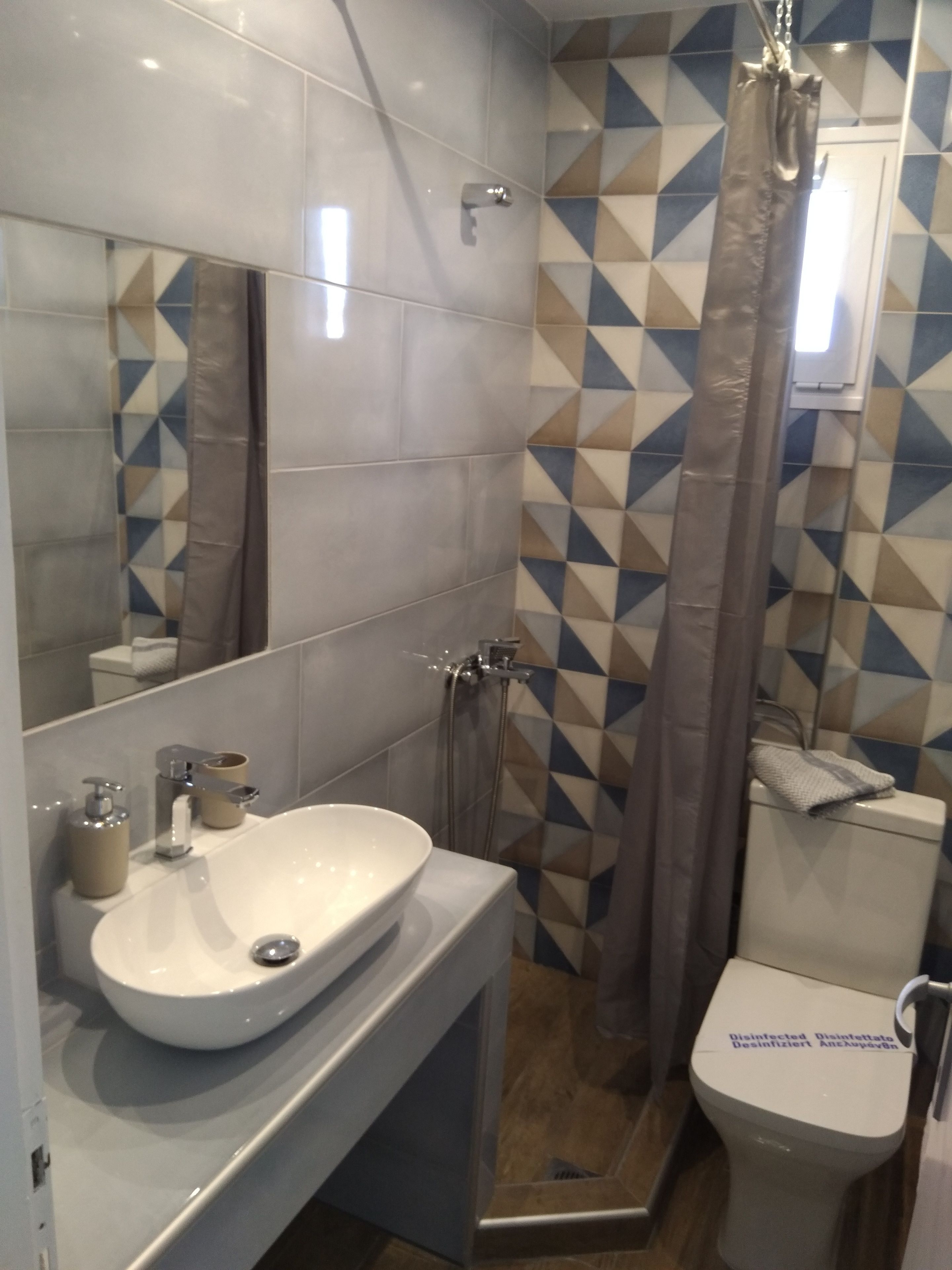 A modern bathroom with a white sink, mirror, and shower. The walls have a geometric tile pattern.