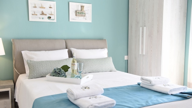A neatly made bed with white and blue bedding, a tray with a bottle and glass, and two towels on the bed. - Dionisos Elia (Nea Propontida)