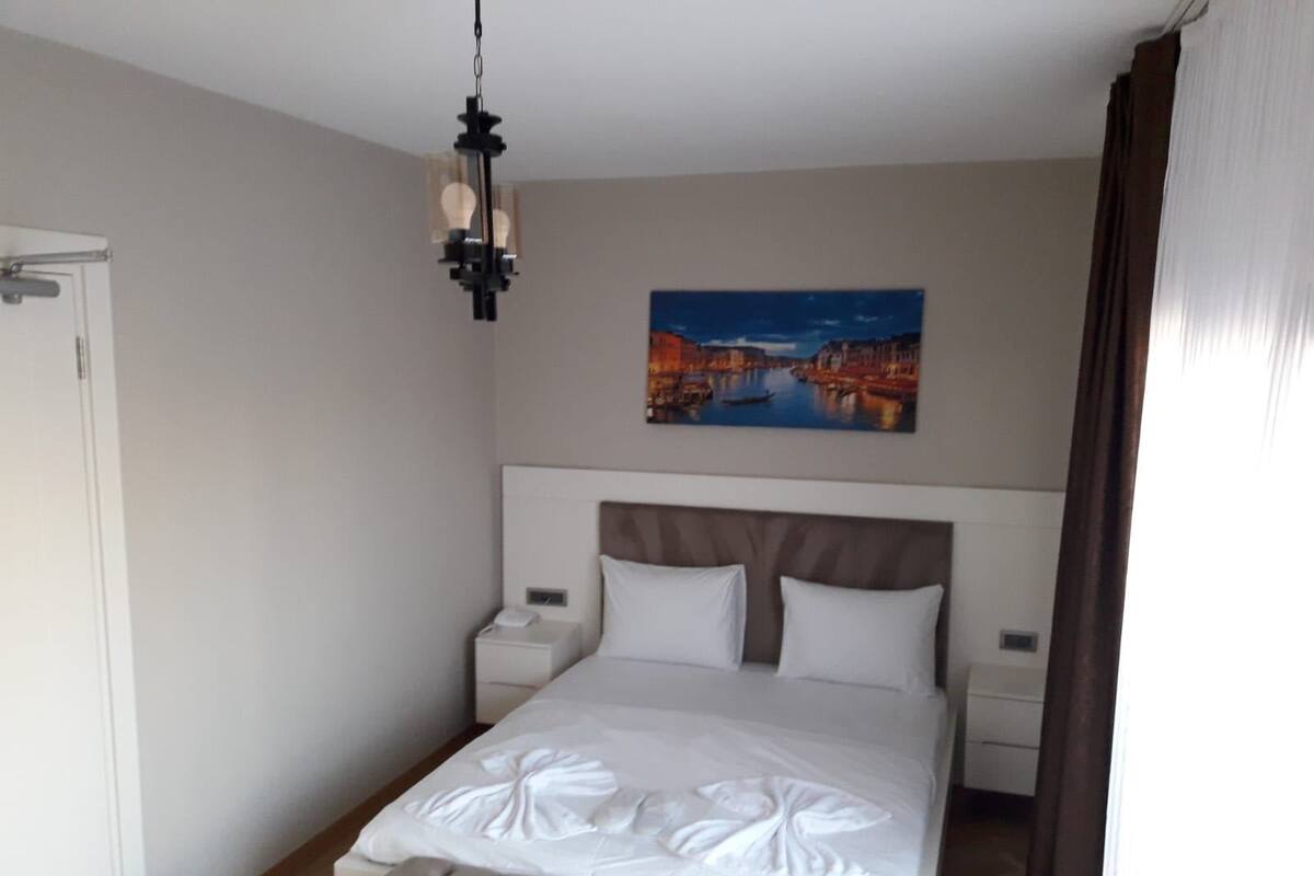 A hotel room with a large bed, a headboard, a nightstand, a bedside table, a hanging light fixture, and a painting of a cityscape.