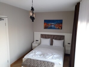 Quarto de hotel com uma cama grande, cabeceira, mesa de cabeceira, luminária pendente e uma pintura de uma paisagem urbana.