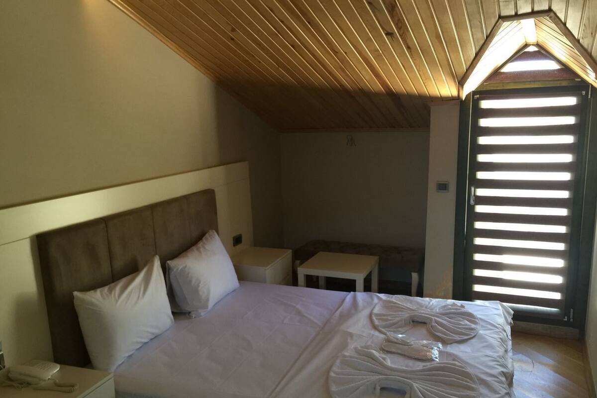 A bedroom with a bed, a bedside table, a small table, and a window with blinds.