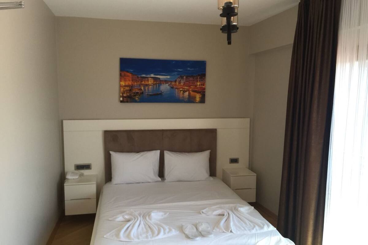 A hotel room with a bed, a nightstand, a bedside table, a painting of a cityscape, and a window with curtains.