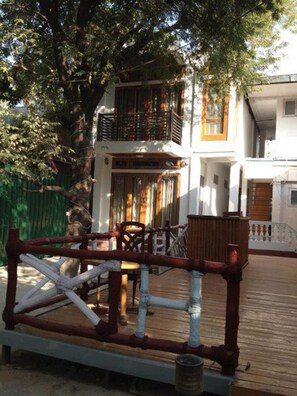 A wooden deck with a railing, a dining table with chairs, and a building with multiple balconies in the background. - Peacock Lodge (Mandalay)