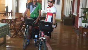 Dos personas en bicicleta en una habitación amplia con suelo de madera, una zona de comedor y una pared roja.