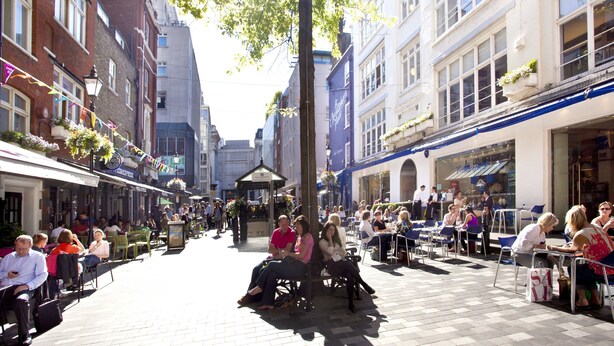 Ein belebtes Straßencafé mit Tischen und Stühlen, umgeben von Gebäuden mit Geschäften und Restaurants.