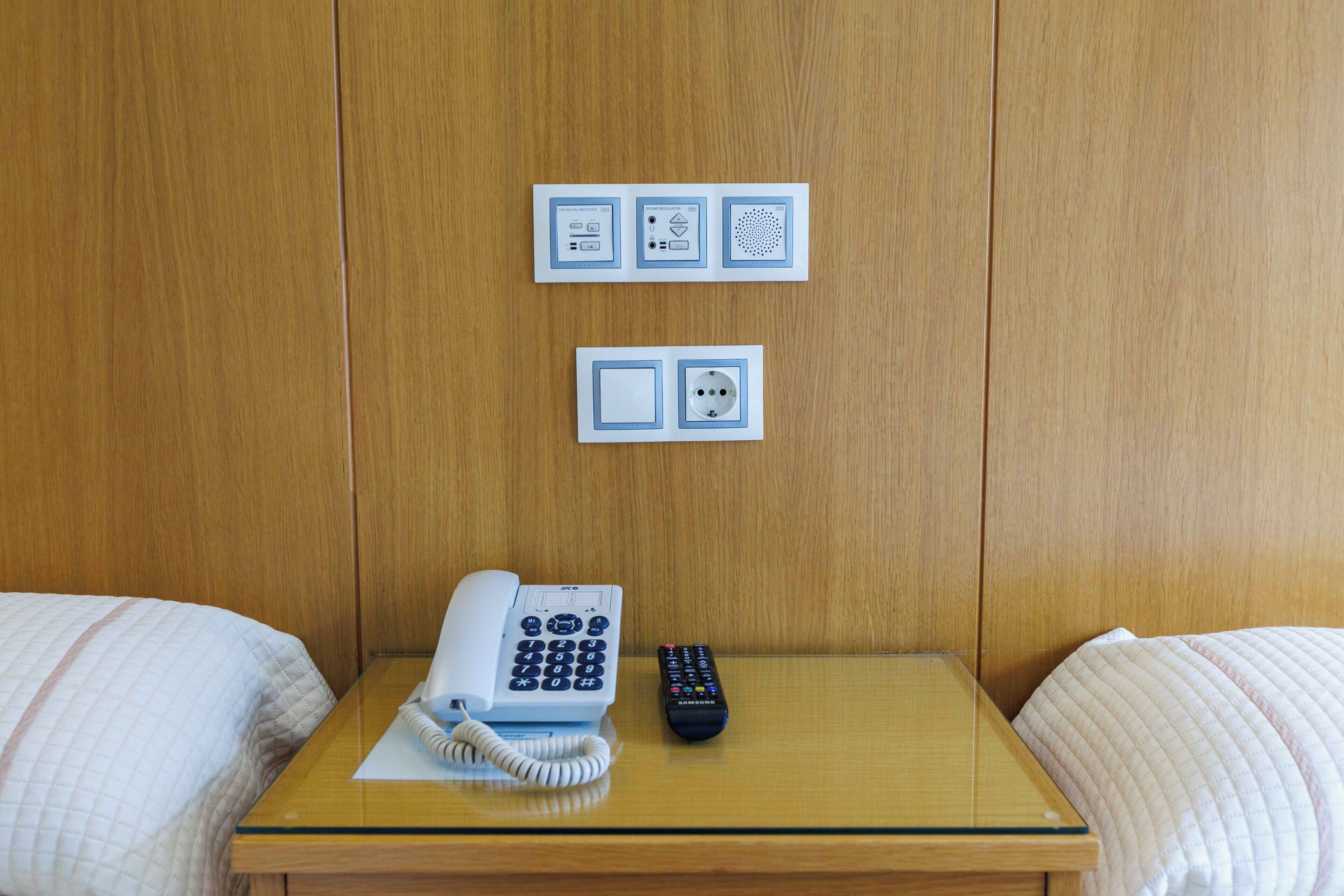 Habitación de hotel con cama, mesita de noche con teléfono y control remoto, y panel eléctrico en la pared.