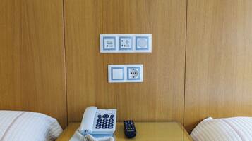 Habitación de hotel con cama, mesita de noche con teléfono y control remoto, y panel eléctrico en la pared.