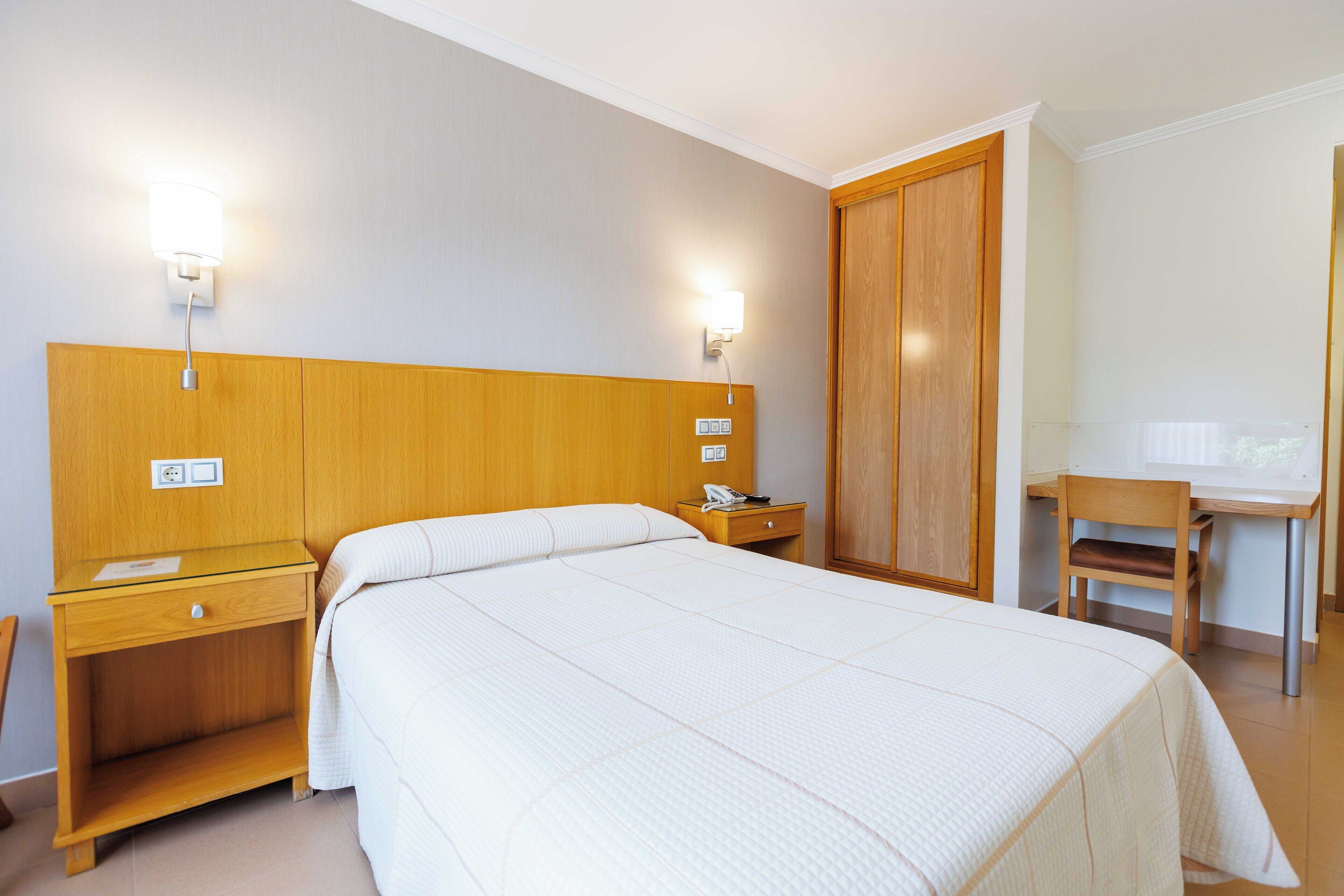 A hotel room with a large bed, a wooden headboard, a desk with a chair, and two wall-mounted lights.