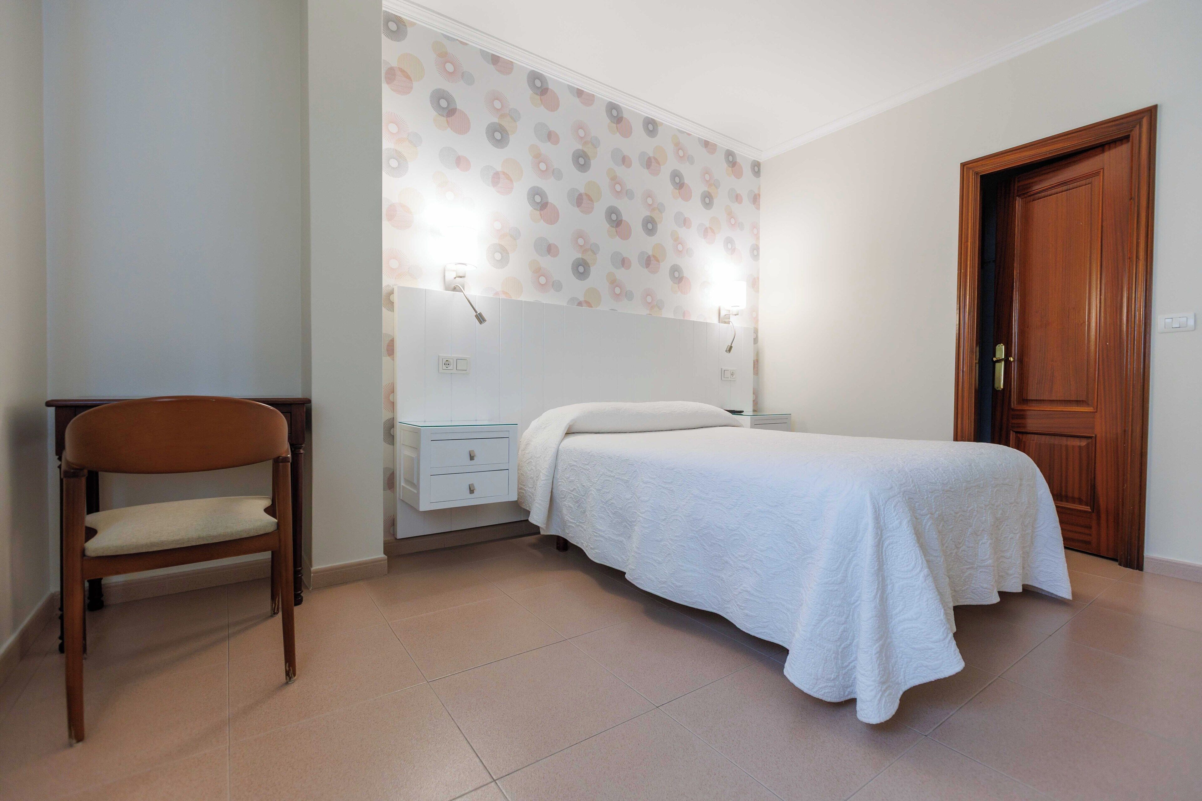 A bedroom with a bed, a chair, a nightstand, a wall-mounted lamp, and a door.