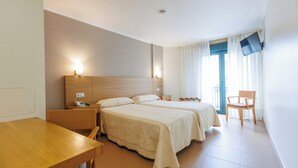 Quarto de hotel com duas camas, uma escrivaninha de madeira, uma cadeira, televisão fixada na parede e uma janela com cortinas.