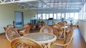 Uma área de estar externa coberta, com mobiliário de vime e mesa de vidro, oferecendo vista da cidade através de grandes janelas.