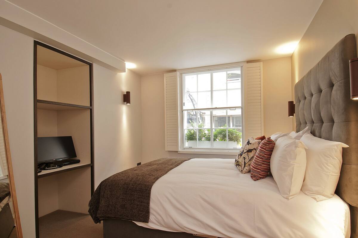 A bedroom with a bed, a television on a shelf, a window, and a headboard with wall sconces.