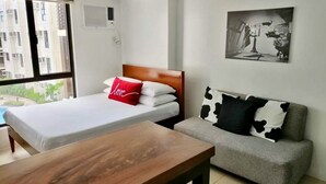 A hotel room with a bed, a sofa, a wooden table, and a window with a view of buildings. - Bella Lifestyle Studio (Tagaytay)