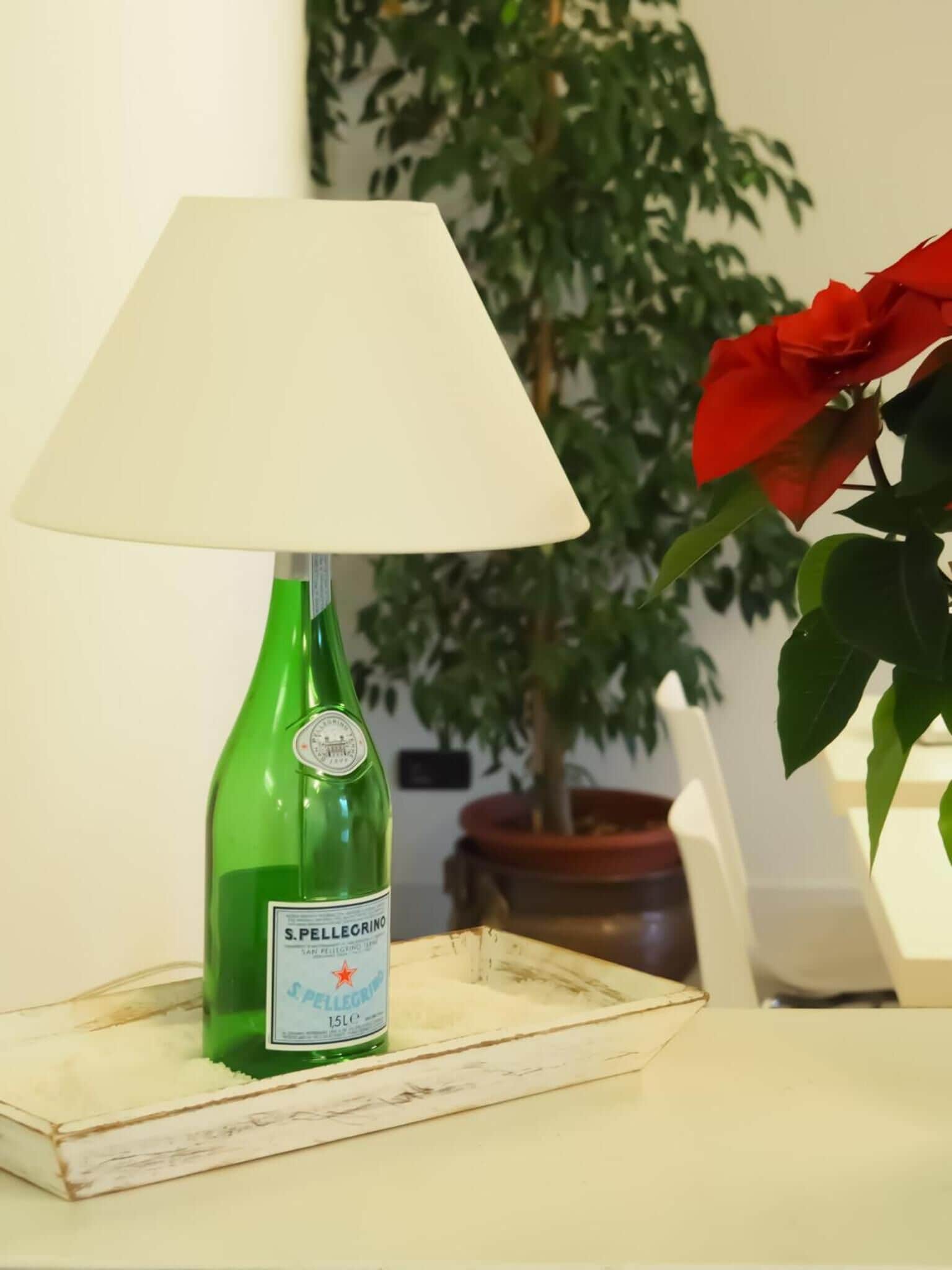 Una bottiglia verde di acqua S. Pellegrino su un vassoio bianco, con un paralume bianco e un fiore rosso sullo sfondo.
