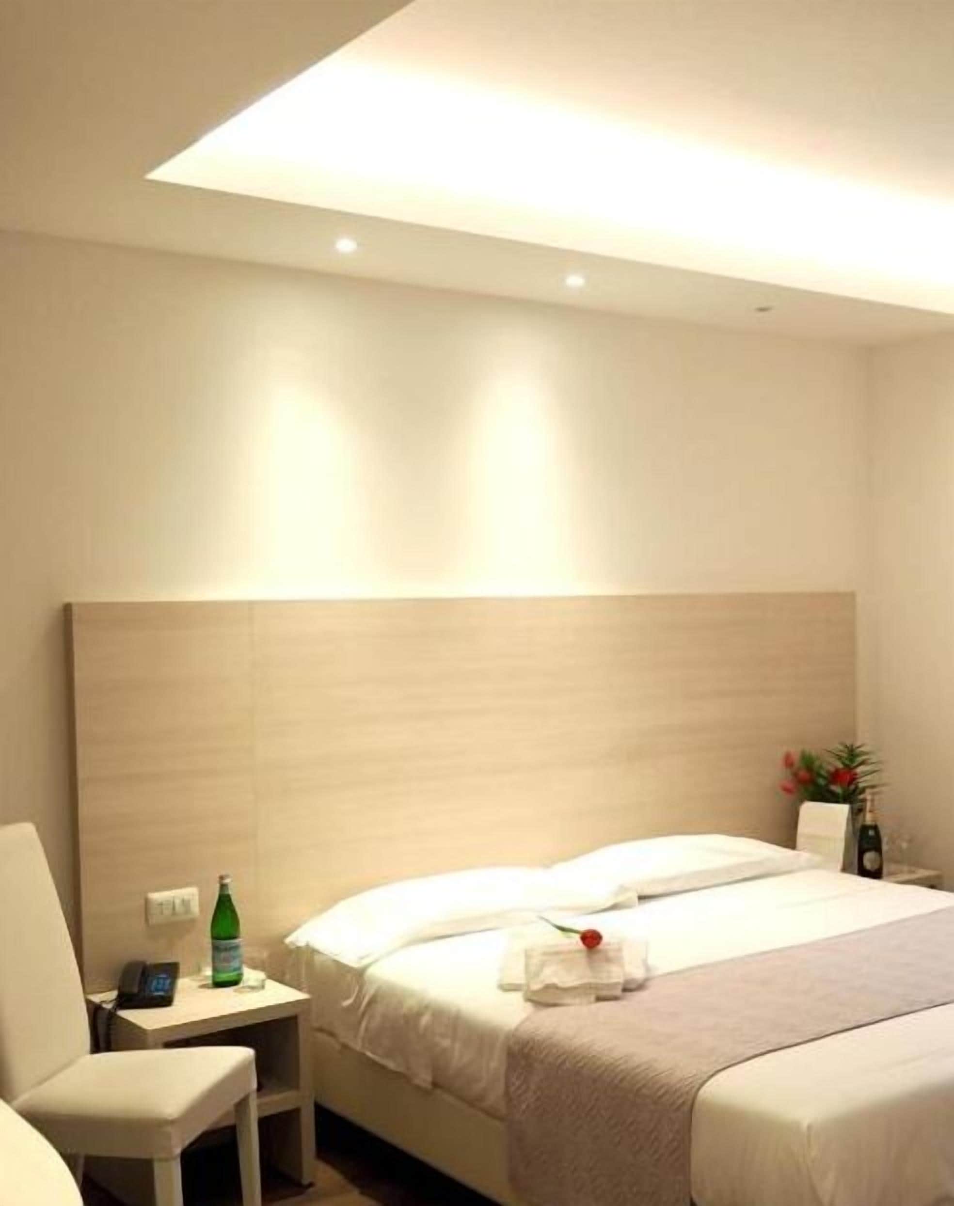 Habitación de hotel con cama, mesita de noche con una botella, una silla y una planta pequeña.