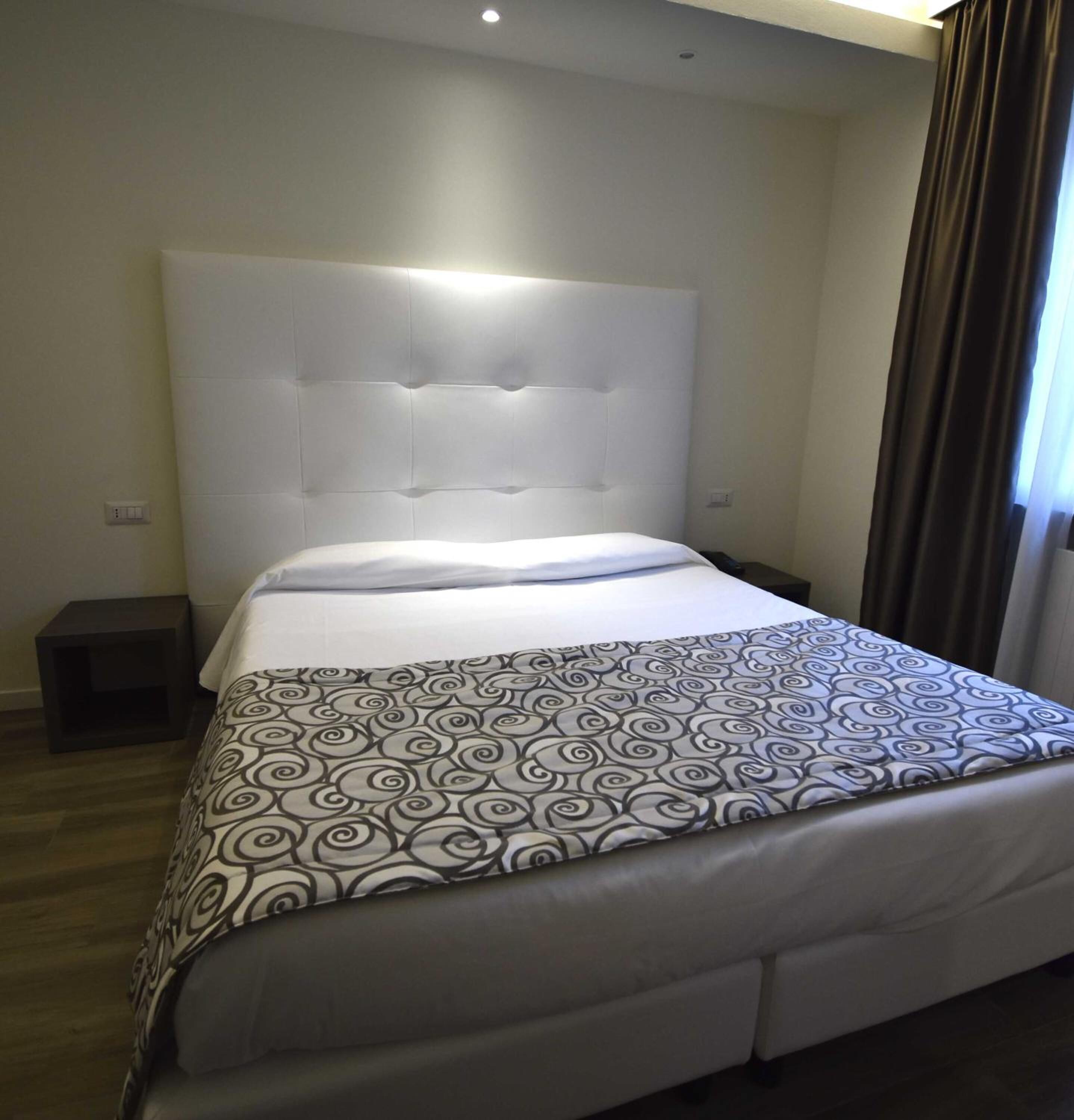 Un letto rifatto con cura, con una coperta a motivi e una testiera imbottita, e un comodino.