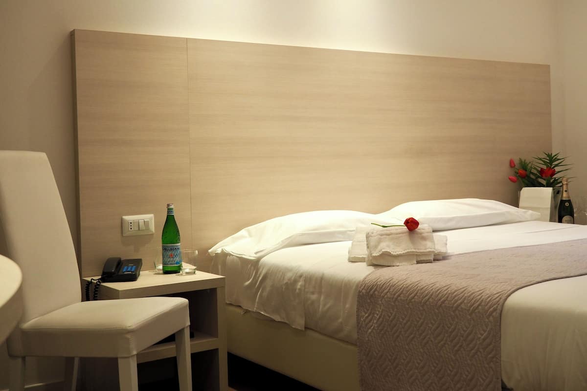 A hotel room with a bed, a bedside table with a bottle, a chair, and a telephone.