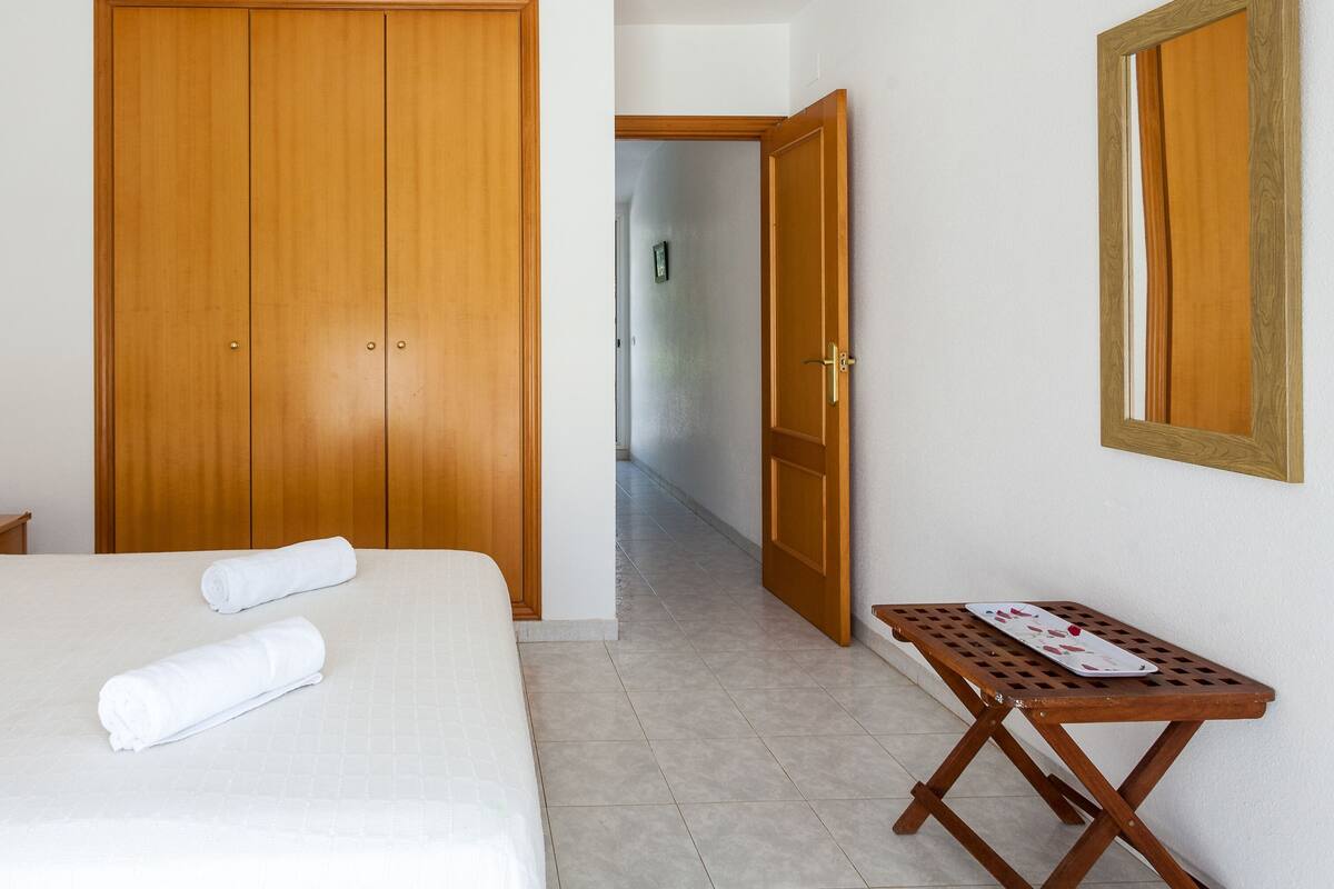 A hotel room with a bed, two rolled towels, a nightstand with a lamp, a wooden wardrobe, and a small table with a menu.