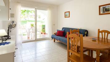Una habitación luminosa con una puerta corrediza de vidrio que da a un jardín, un sofá azul con cojines rojos, una mesa de comedor de madera y un ventilador a la izquierda.