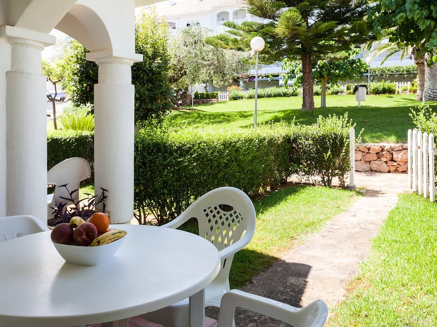 A white table with a bowl of fruit and two white chairs in a sunny outdoor area with a well-maintained lawn and a stone wall in the background.