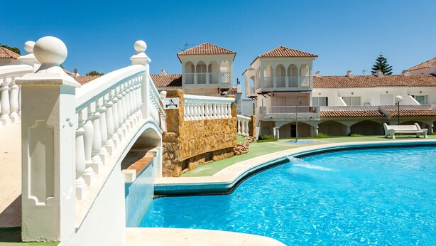 A swimming pool with a stone wall and white balustrade, surrounded by buildings with tiled roofs and balconies. - Al-andalus Casa Azahar (Alcala de Xivert)