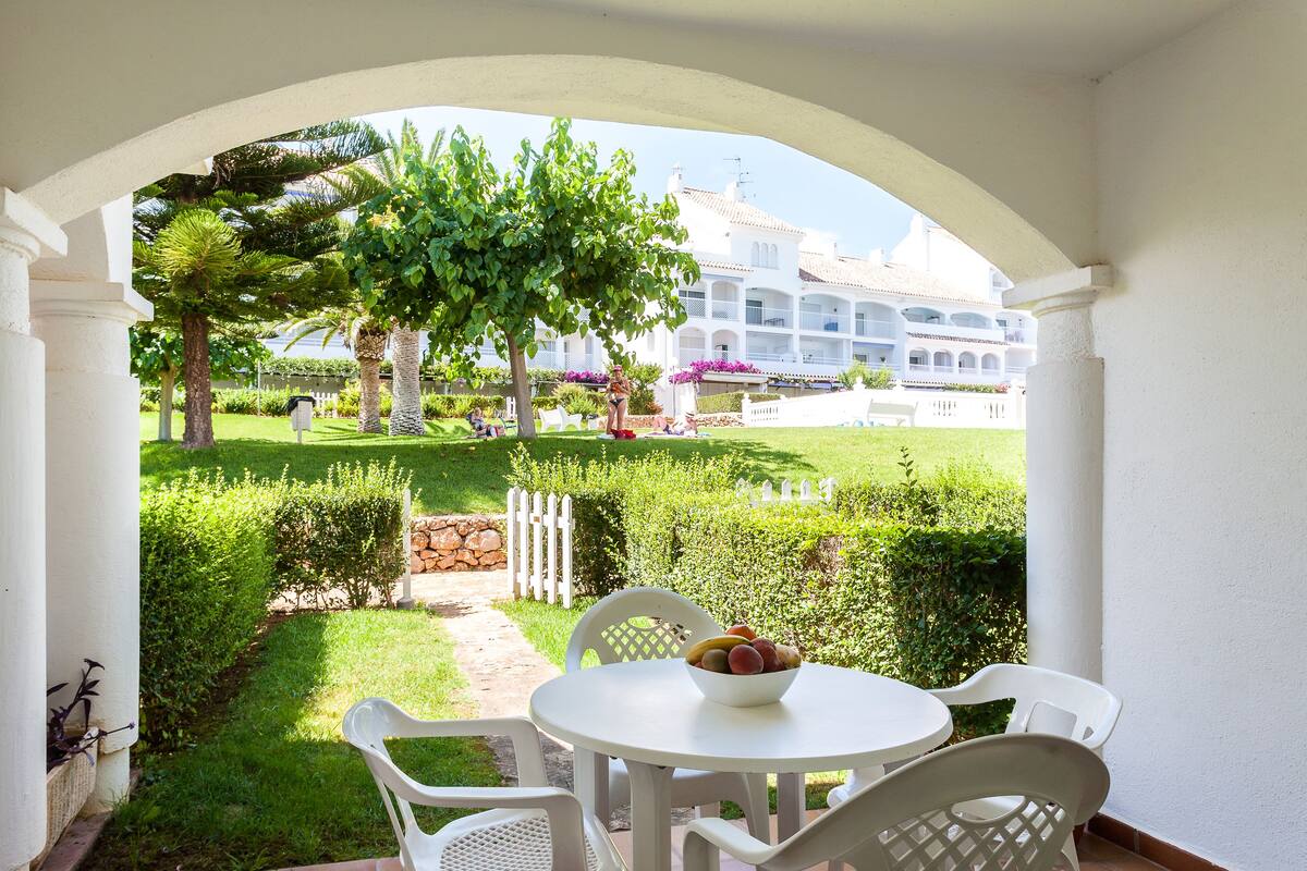 A patio with a white table and chairs, a bowl of fruit, and a view of a well-maintained garden and building in the background.