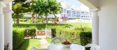 Une terrasse avec une table et des chaises blanches, un saladier rempli de fruits, et une vue sur un jardin bien entretenu et un bâtiment en arrière-plan.