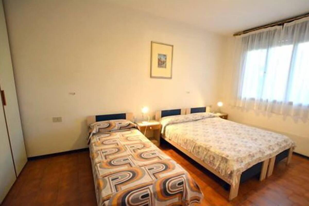 Ein Schlafzimmer mit zwei Betten, einem Nachttisch mit Lampe, einem gerahmten Bild an der Wand und einem Fenster mit Vorhängen.