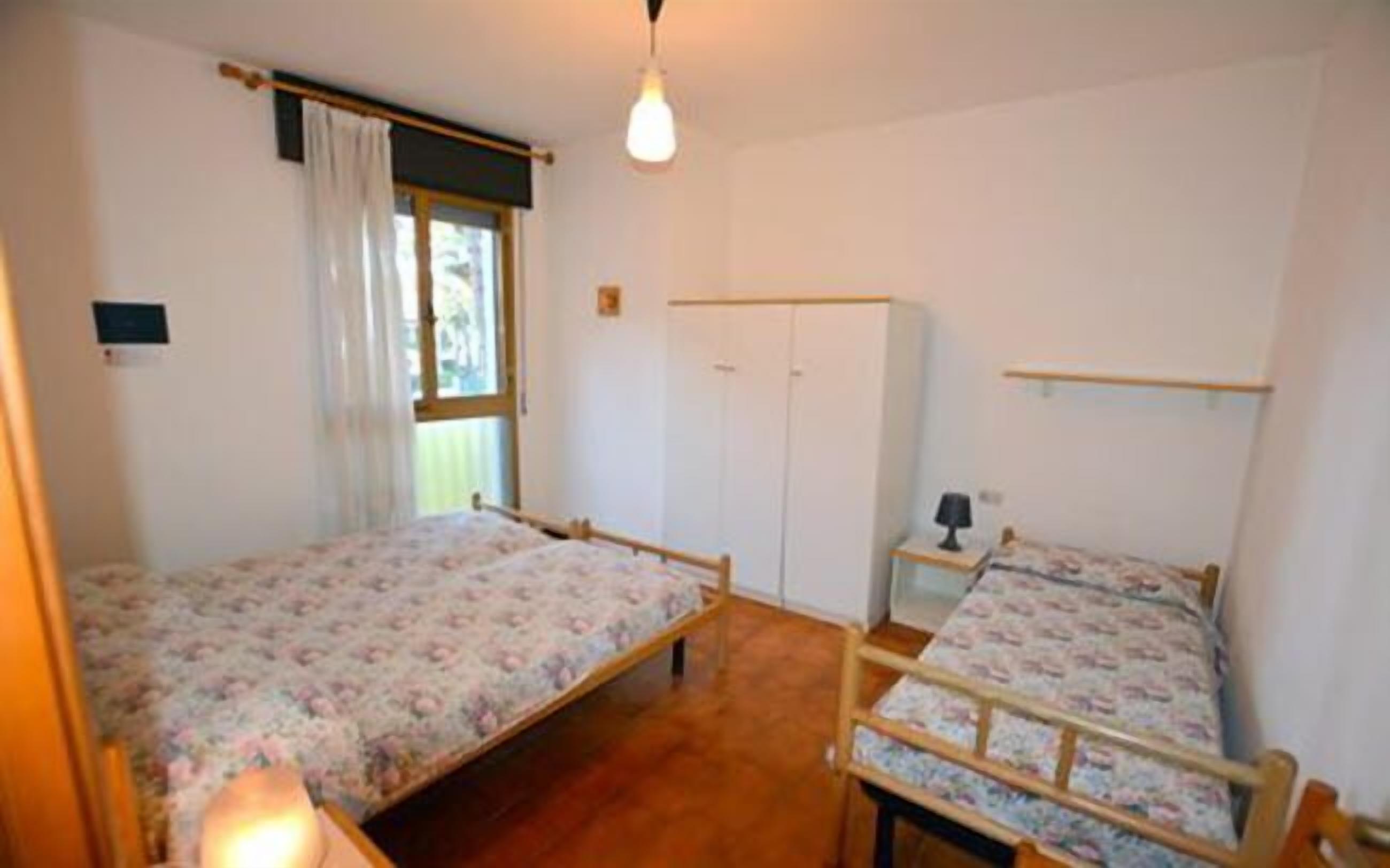 Quarto com duas camas de solteiro, um guarda-roupa, mesa de cabeceira com abajur e janela com cortinas.