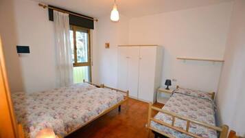 Quarto com duas camas de solteiro, um guarda-roupa, mesa de cabeceira com abajur e janela com cortinas.