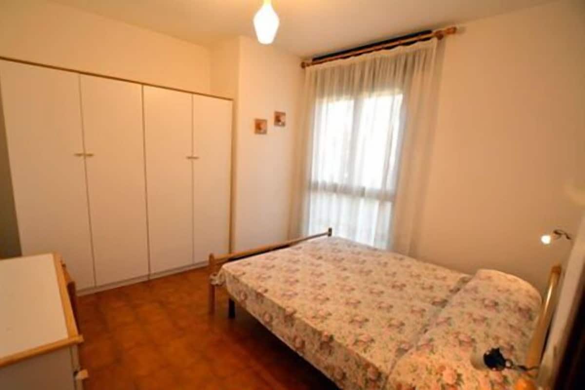 Ein Schlafzimmer mit Bett, Kleiderschrank, einem kleinen Tisch und einem Fenster mit Vorhängen.