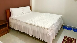A single bed with a white mattress and matching bedding, a wooden headboard, a small wooden nightstand, and a wall-mounted shelf with rolled-up towels. - IQ Hotel (Ho Chi Minh City)