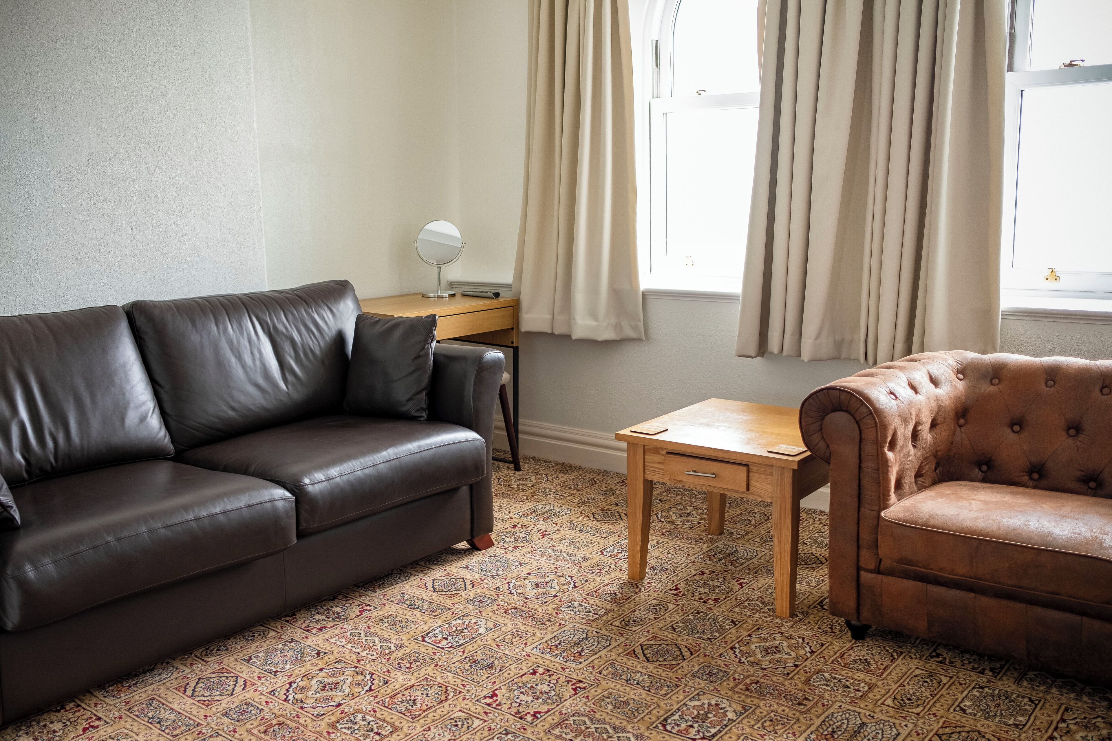 Una habitación con un sofá de cuero marrón, un sillón marrón más pequeño, una mesita de madera y una alfombra estampada.