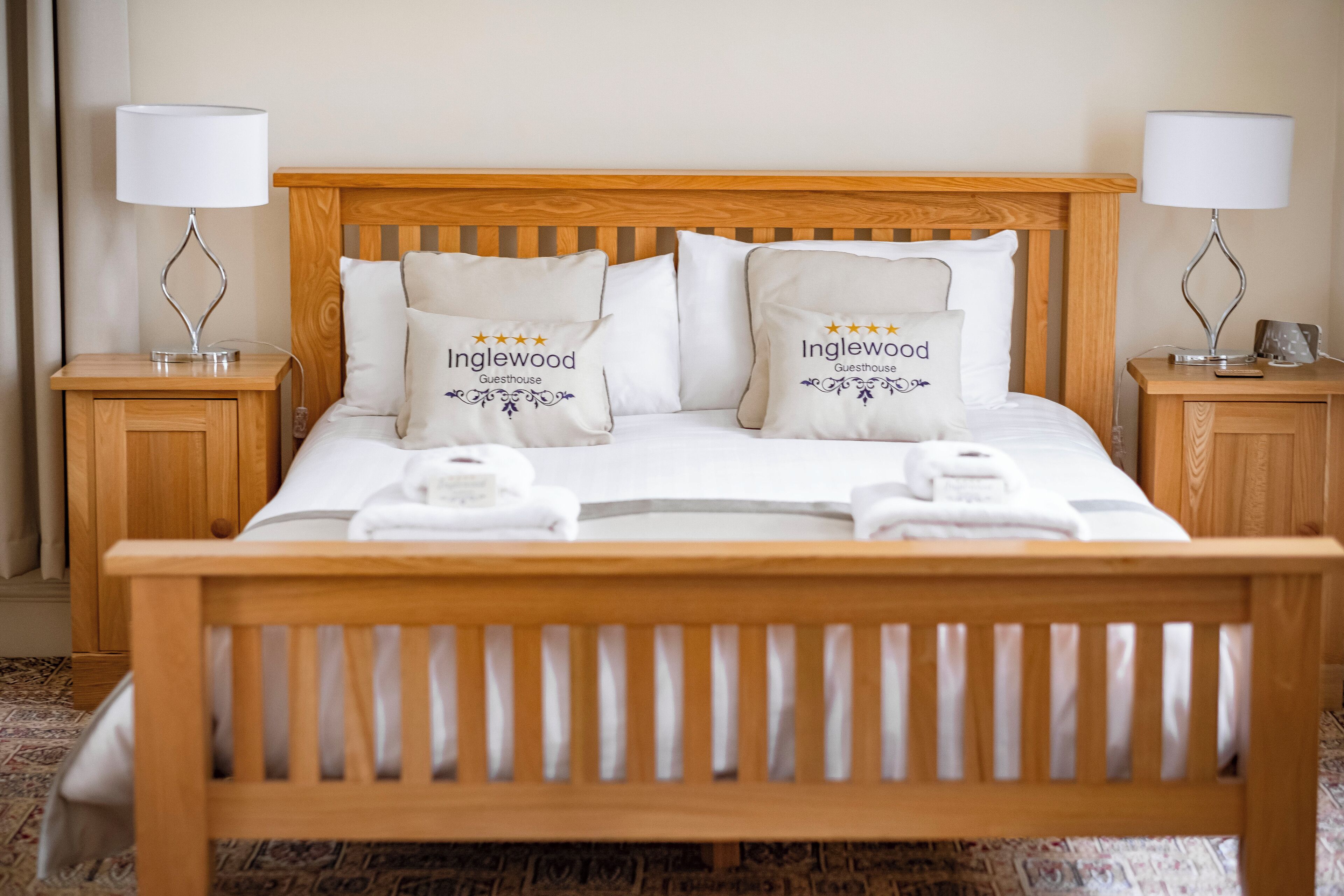 A neatly made bed with two pillows labeled "Inglewood Guesthouse," a wooden headboard, and two bedside tables with lamps.