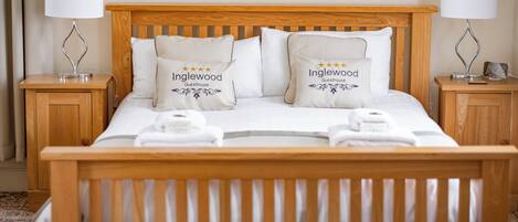 A neatly made bed with two pillows labeled "Inglewood Guesthouse," a wooden headboard, and two bedside tables with lamps.