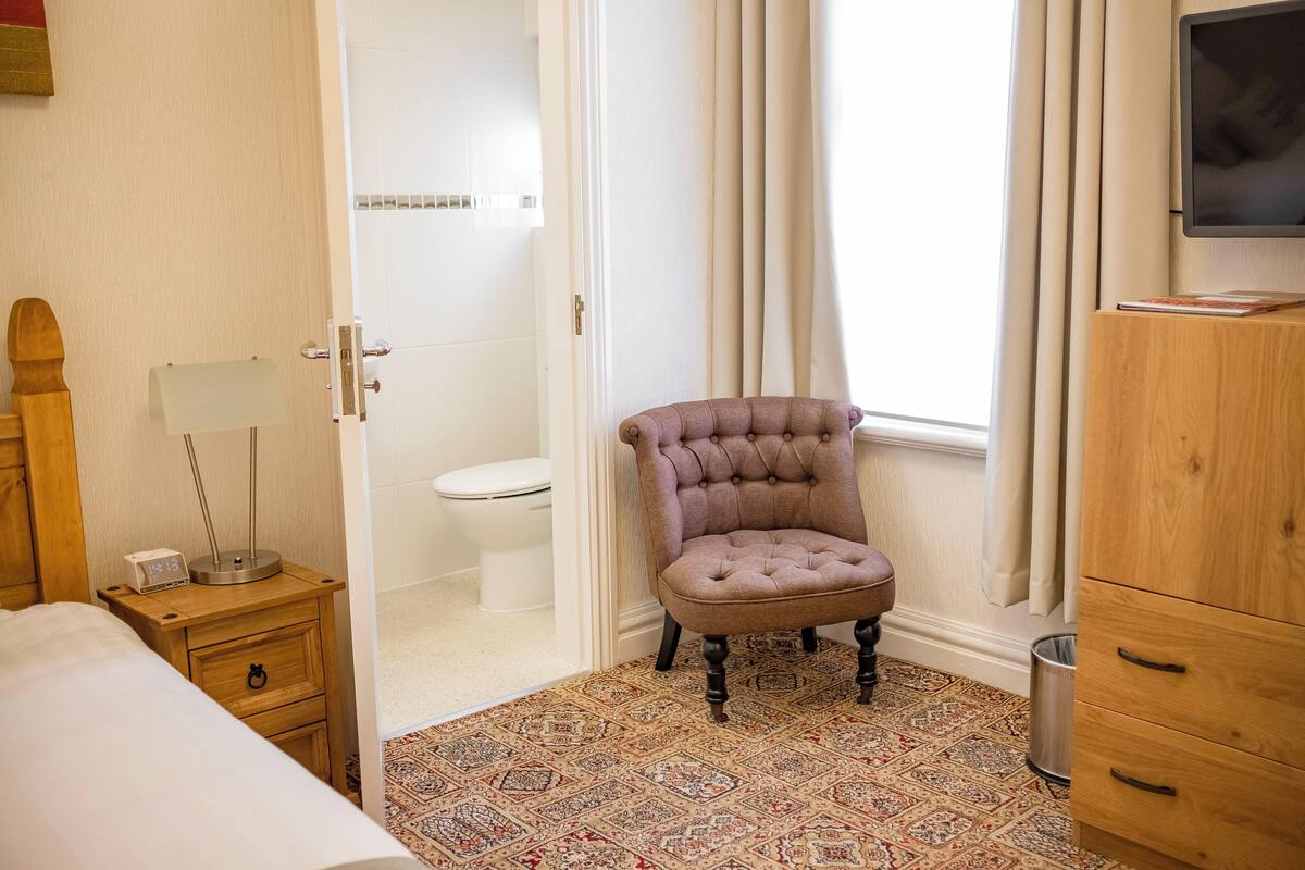 A hotel room with a bed, a wooden nightstand with a lamp, a tufted armchair, a wooden dresser, and a television on the wall.