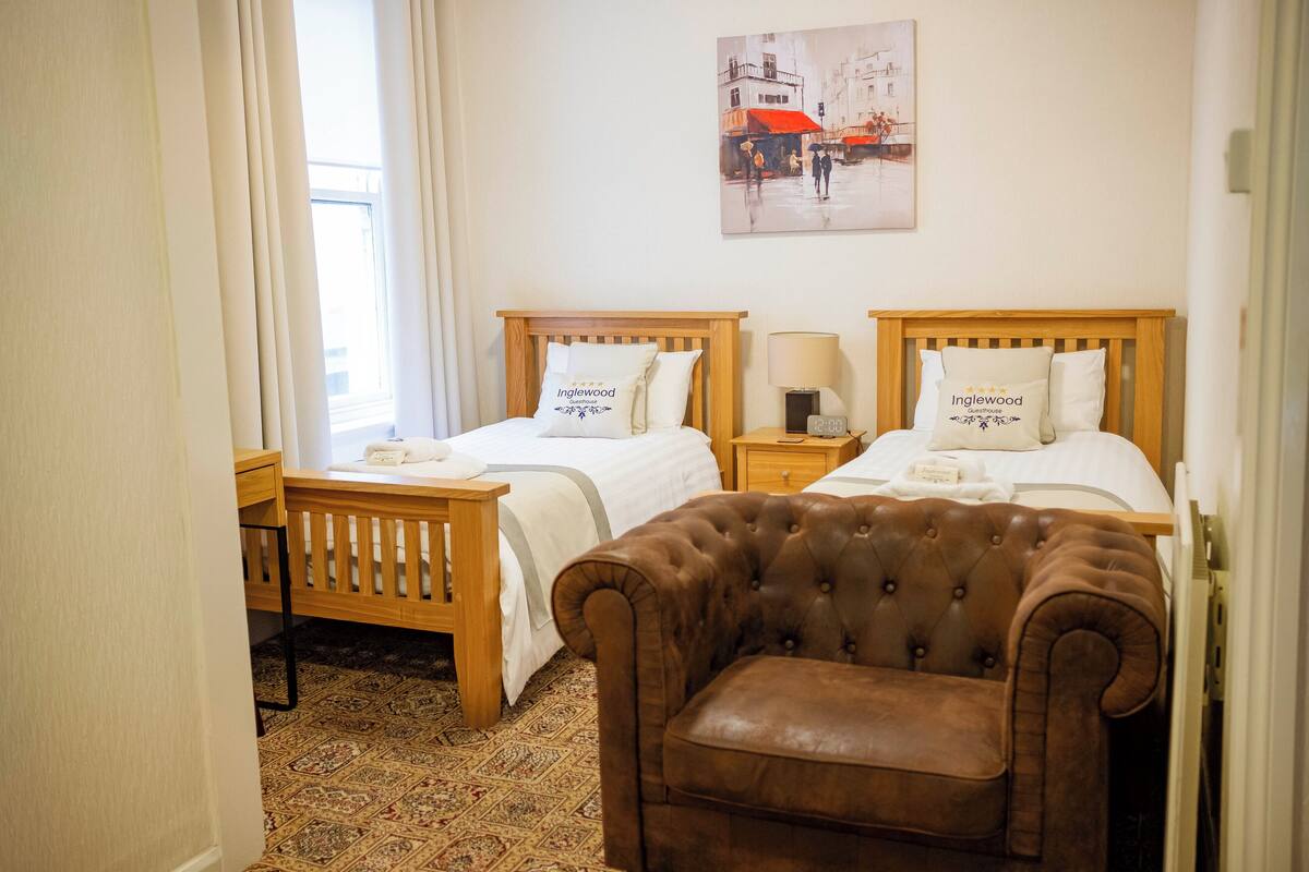 A hotel room with two beds, a wooden headboard, a brown leather armchair, a patterned carpet, and a painting on the wall.