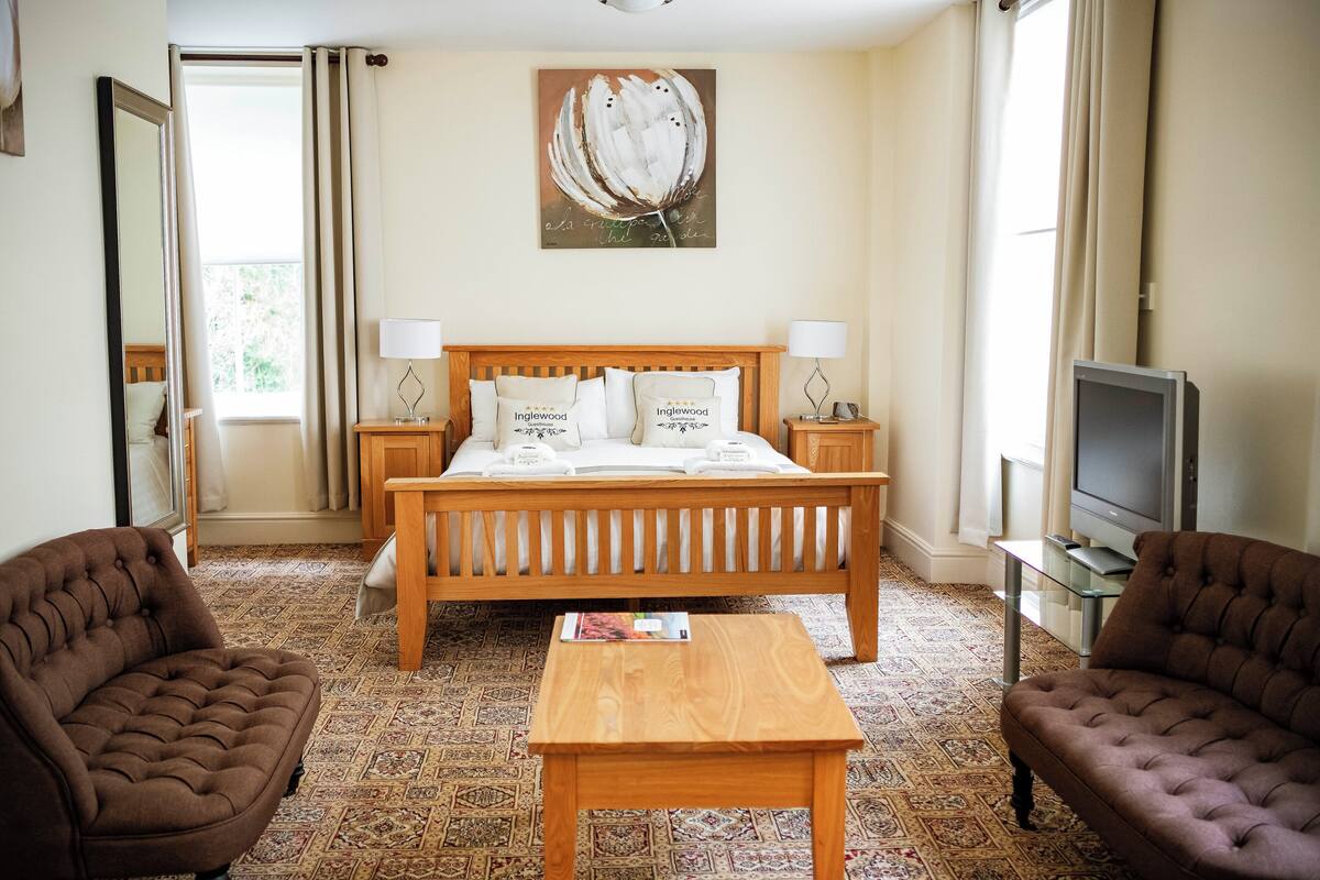 A hotel room with a wooden bed, two armchairs, a small table, a TV, and a painting on the wall.