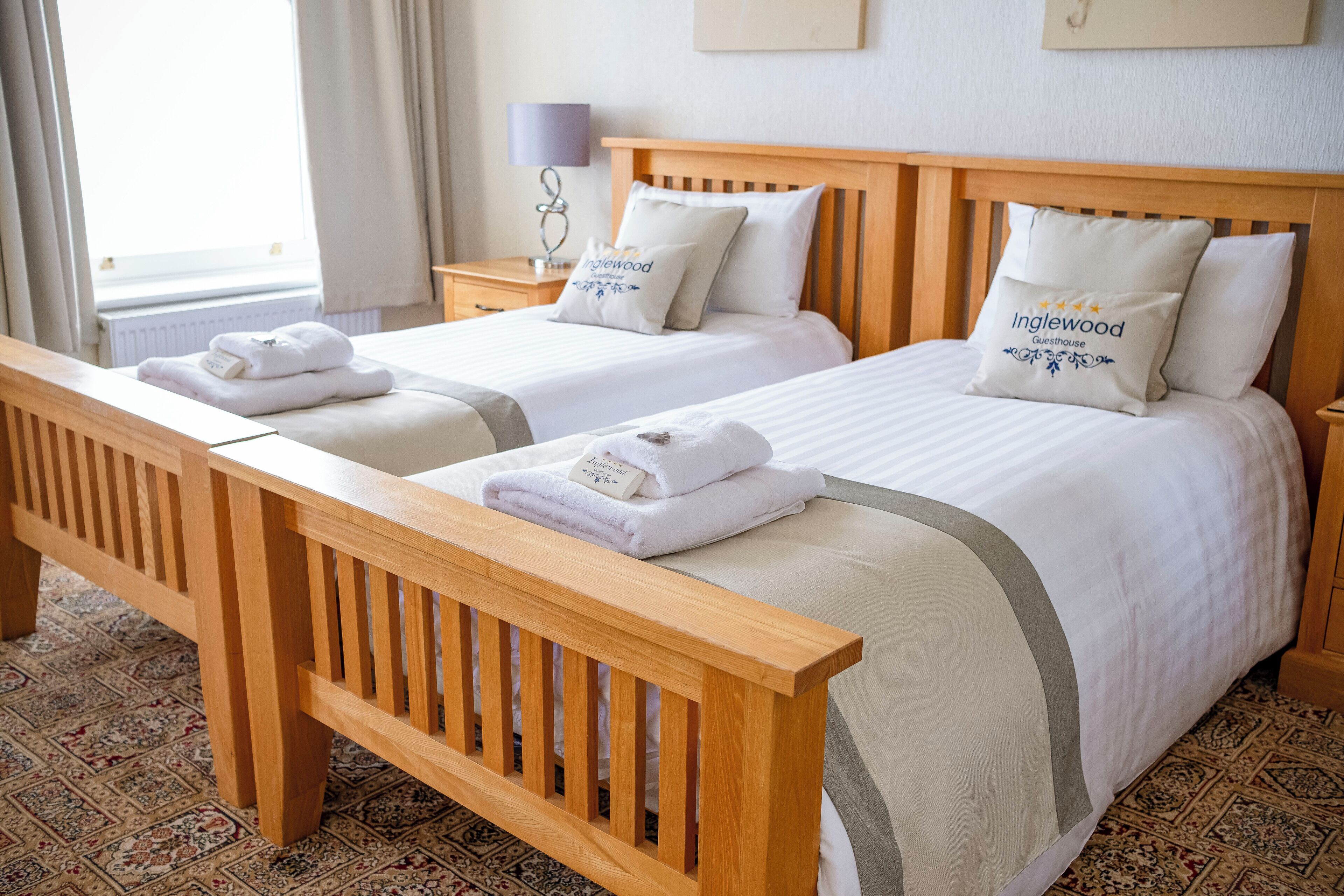 A hotel room with two beds, a wooden bench, a nightstand with a lamp, and a window with curtains.