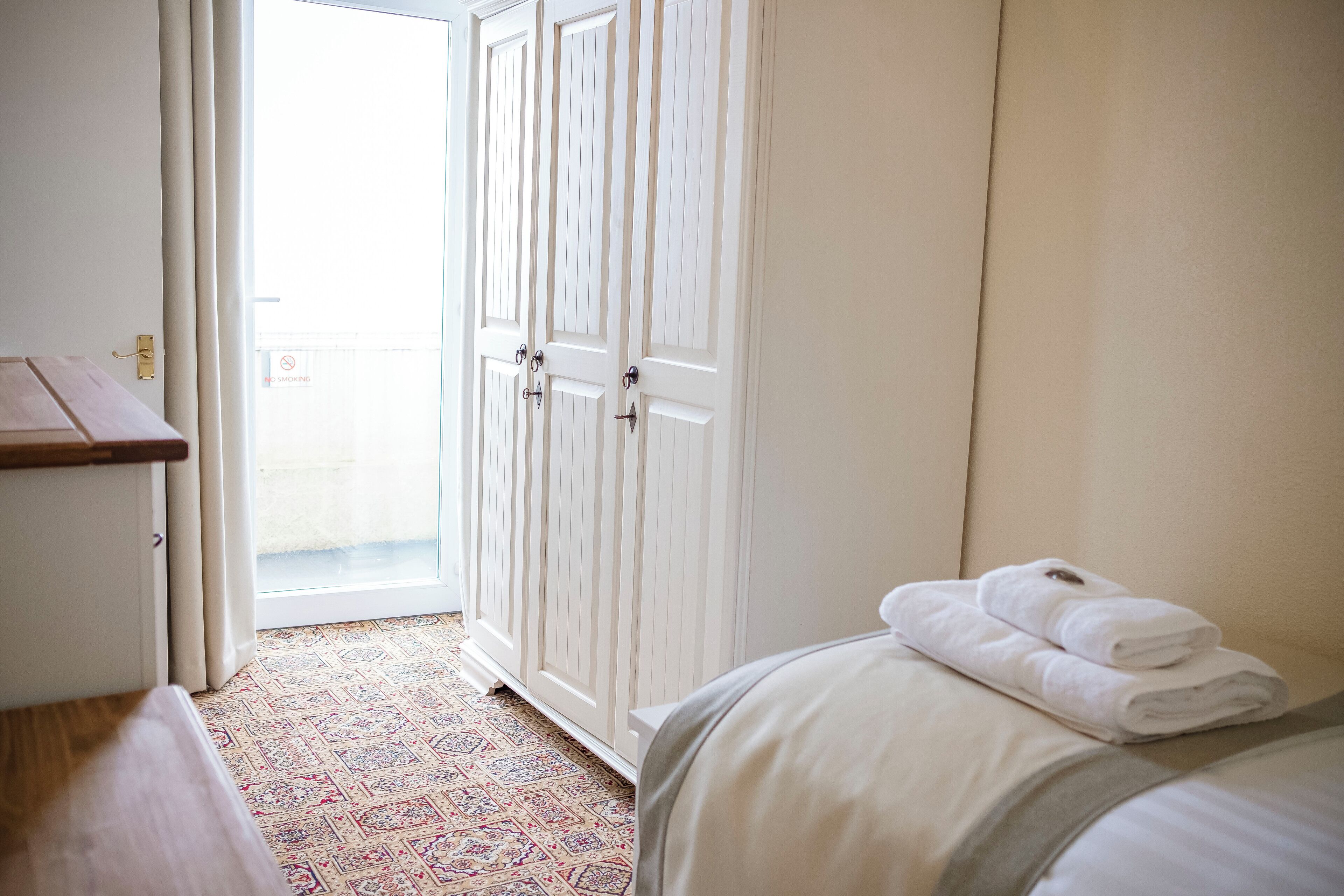 A hotel room with a bed, a wooden bench, a patterned rug, and a set of white doors leading to a balcony.
