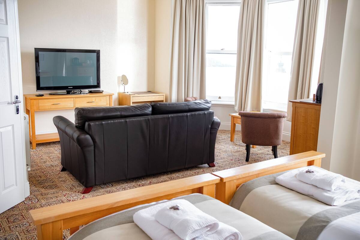 A hotel room with a black leather sofa, a wooden TV stand, a small wooden table, a brown chair, and two beds with white towels.