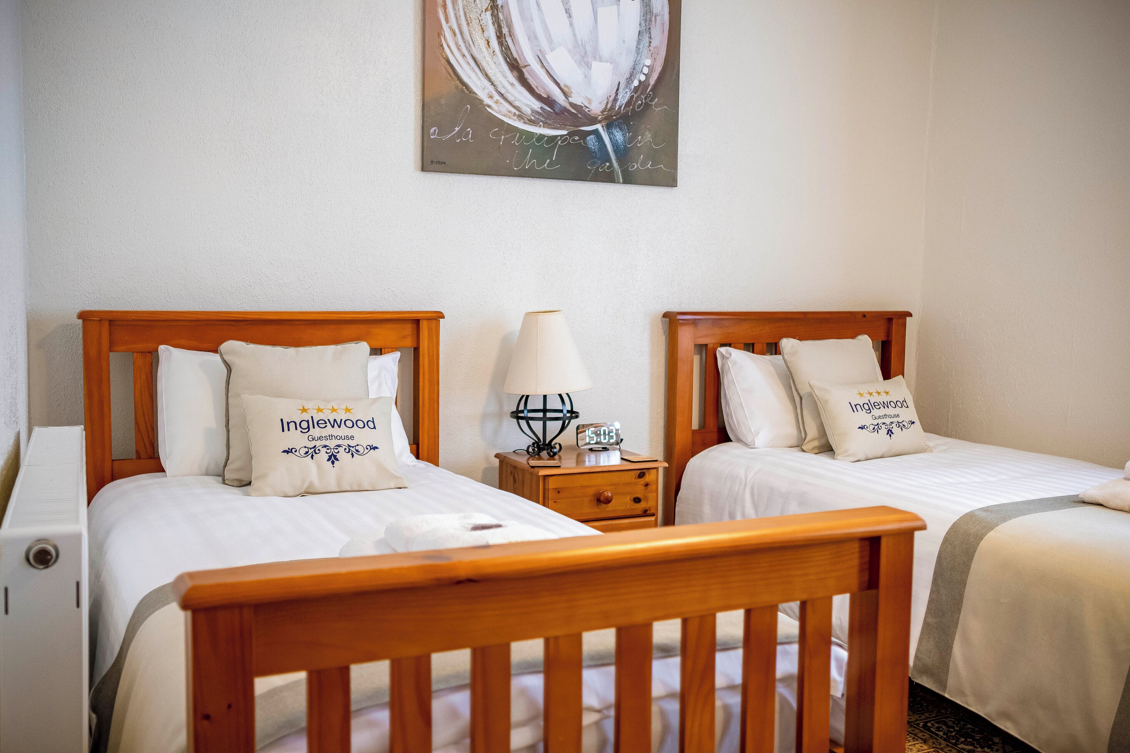 A hotel room with two beds, each with a wooden headboard, white bedding, and a pillow labeled "Inglewood Guesthouse". There is a nightstand with a lamp and a clock between the beds.
