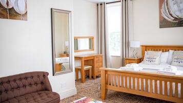 A hotel room with a bed, a wooden desk, a brown sofa, a mirror, and artwork on the walls.