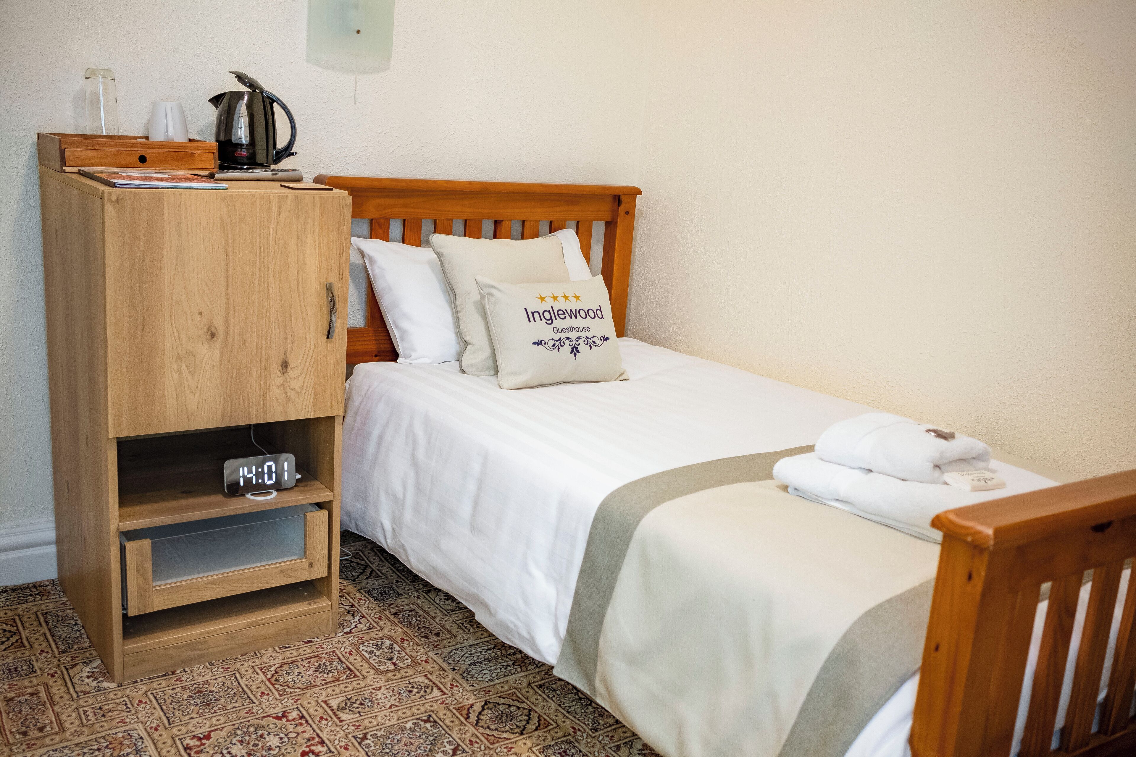 A neatly made bed with a pillow labeled "Inglewood Guesthouse," a wooden nightstand with a clock, and a coffee maker.