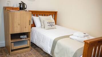 A neatly made bed with a pillow labeled "Inglewood Guesthouse," a wooden nightstand with a clock, and a coffee maker.