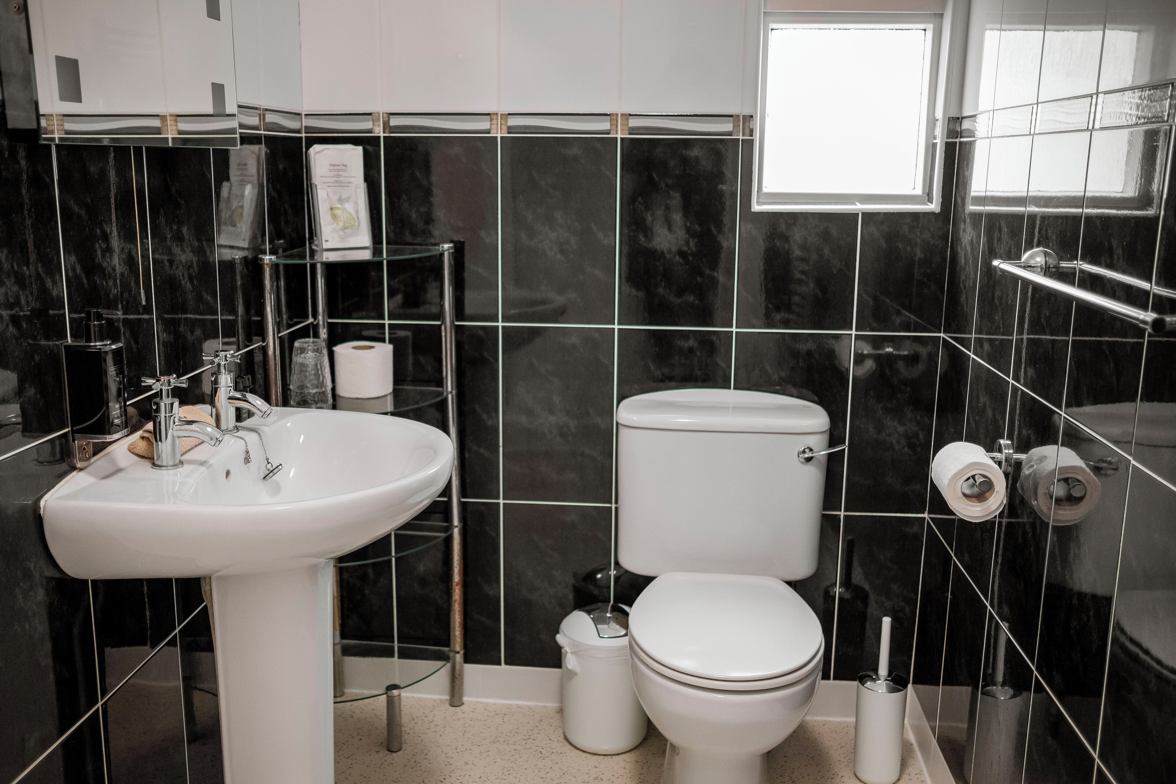 A modern bathroom with a white sink, toilet, and bidet, black tiled walls, and a window providing natural light.